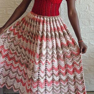 Op de afbeelding: Een handgemaakte, rode gehaakte top met korte mouwen en een sweetheart halslijn. De rok heeft een chevronpatroon in tinten roze, wit en bruin. De rok is een lange, vloeiende stijl, perfect voor een zomerdag.