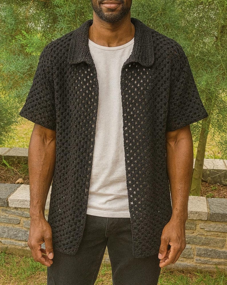 Puede incluir: Una camisa negra de crochet de tejido abierto, con mangas cortas y cuello. La camisa se lleva sobre una camiseta gris claro y se combina con vaqueros oscuros. El fondo presenta follaje verde y un muro de piedra.