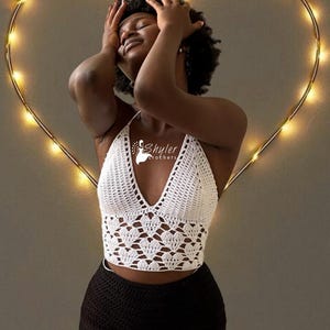 May include: A white crochet halter top with a deep V-neck and a heart-shaped pattern on the bodice. The model is wearing black shorts with orange and white trim. The background features a heart-shaped string of lights.
