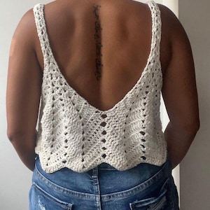 May include: Cream-colored crochet top with a deep V-neck and wide straps. The top has a scalloped hemline and a textured, open weave design. The person is wearing blue denim jeans.