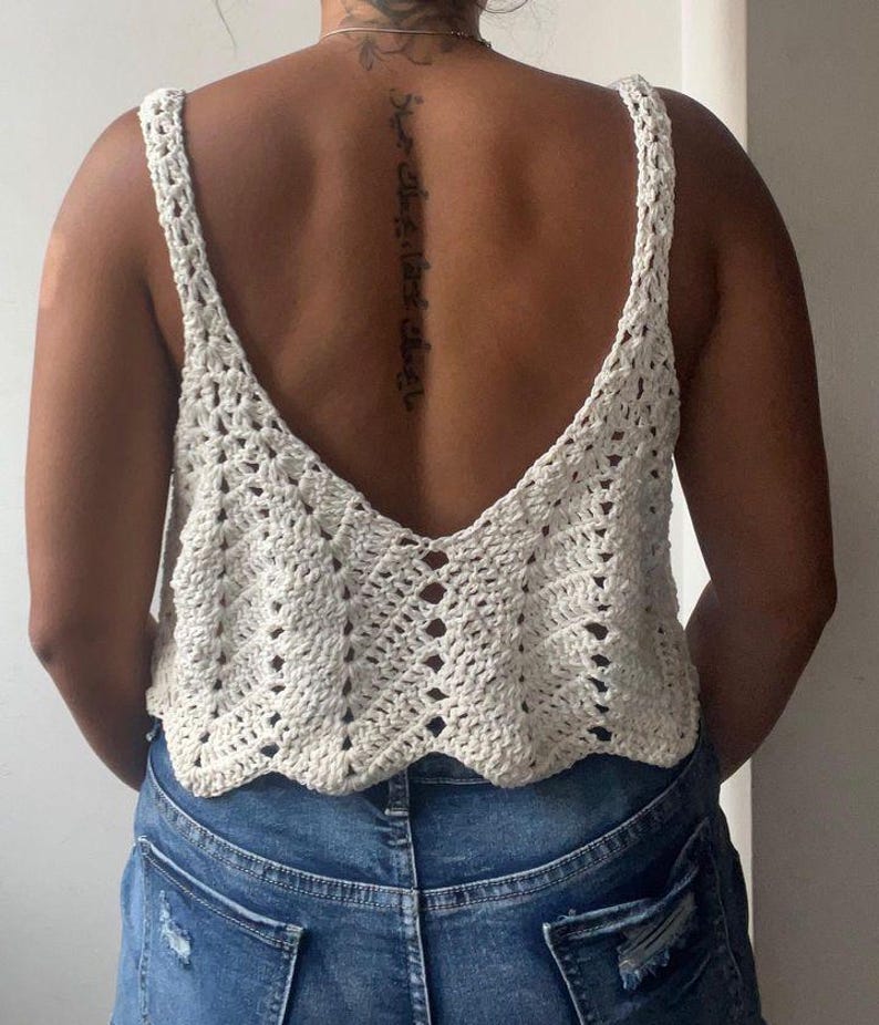 May include: Cream-colored crochet top with a deep V-neck and wide straps. The top has a scalloped hemline and a textured, open weave design. The person is wearing blue denim jeans.