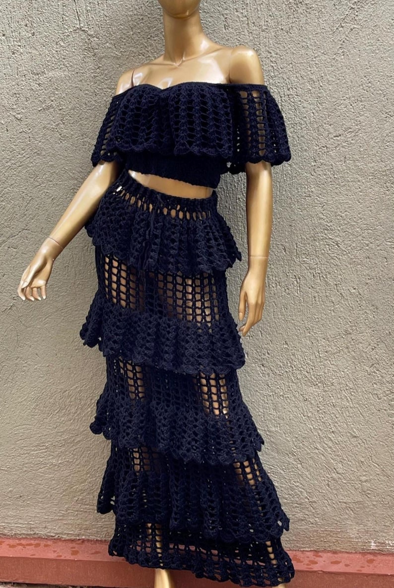 Peut inclure: Un ensemble deux pi&egrave;ces en crochet noir compos&eacute; d'un haut court &agrave; &eacute;paules d&eacute;nud&eacute;es avec des manches &agrave; volants et d'une jupe longue assortie &agrave; &eacute;tages. Le v&ecirc;tement pr&eacute;sente un motif en maille ajour&eacute;e cr&eacute;ant un aspect textur&eacute; semi-transparent.