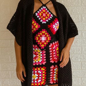 Puede incluir: Un c&aacute;rdigan de crochet negro y un conjunto a juego de top halter y falda. El conjunto presenta un colorido patr&oacute;n de cuadrados de la abuela en rosa, amarillo y blanco sobre un fondo negro. El c&aacute;rdigan tiene mangas cortas.