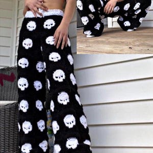May include: Black crochet pants with a white skull pattern. The pants have a wide leg design and a white waistband. The skulls are evenly spaced across the fabric. The pants are worn with black and white sneakers.
