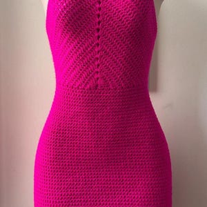 Puede incluir: Vestido de ganchillo fucsia vibrante exhibido en un maniquí. La parte superior de estilo halter se transforma en un corpiño ajustado y una falda recta. El vestido está hecho de un hilo texturizado, mostrando un patrón de puntada detallado. El vestido es de color sólido.