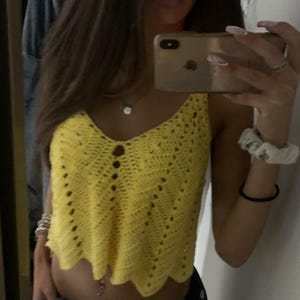 May include: A yellow crochet top with a V-neckline and scalloped hem. The top has a textured, open-weave design with small button details. The garment is sleeveless and appears to be handmade.