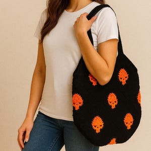 May include: Black crochet tote bag with orange skull designs. The bag has a rounded shape and long straps. The skulls are evenly spaced across the front of the bag. The bag is being held by a person wearing a white t-shirt and blue jeans.