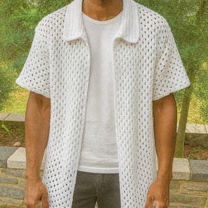Puede incluir: Una camisa blanca de crochet de manga corta con cuello. La camisa tiene la parte delantera abierta, revelando una camiseta blanca debajo. El dise&ntilde;o de crochet presenta un patr&oacute;n de rejilla. La persona lleva vaqueros grises oscuros.