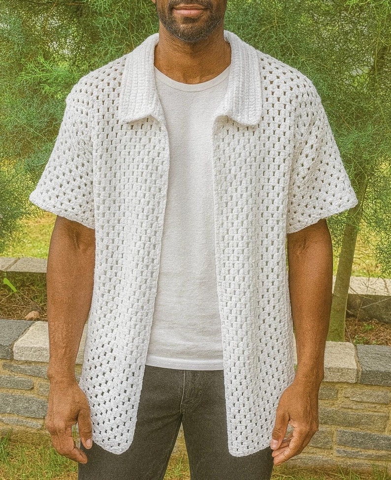 Puede incluir: Una camisa blanca de crochet de manga corta con cuello. La camisa tiene la parte delantera abierta, revelando una camiseta blanca debajo. El dise&ntilde;o de crochet presenta un patr&oacute;n de rejilla. La persona lleva vaqueros grises oscuros.