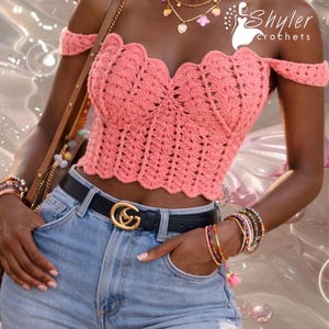 Op de afbeelding: Een koraalkleurige, off-the-shoulder gehaakte top met een geschulpte halslijn. De top wordt gecombineerd met een lichtblauwe spijkerbroek, een zwarte riem met een gouden gesp en gelaagde kettingen en armbanden. De afbeelding bevat de tekst "Shyler crochets".