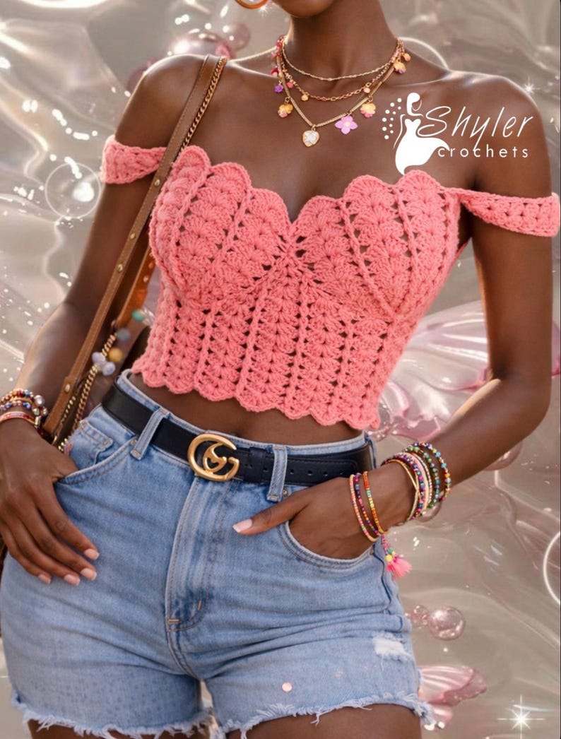 Op de afbeelding: Een koraalkleurige, off-the-shoulder gehaakte top met een geschulpte halslijn. De top wordt gecombineerd met een lichtblauwe spijkerbroek, een zwarte riem met een gouden gesp en gelaagde kettingen en armbanden. De afbeelding bevat de tekst "Shyler crochets".
