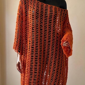 Puede incluir: Vestido túnica de crochet naranja con tejido abierto, escote asimétrico y mangas largas. El vestido se exhibe en un maniquí negro, mostrando su diseño suelto y fluido. El patrón de crochet crea un aspecto texturizado y aireado.