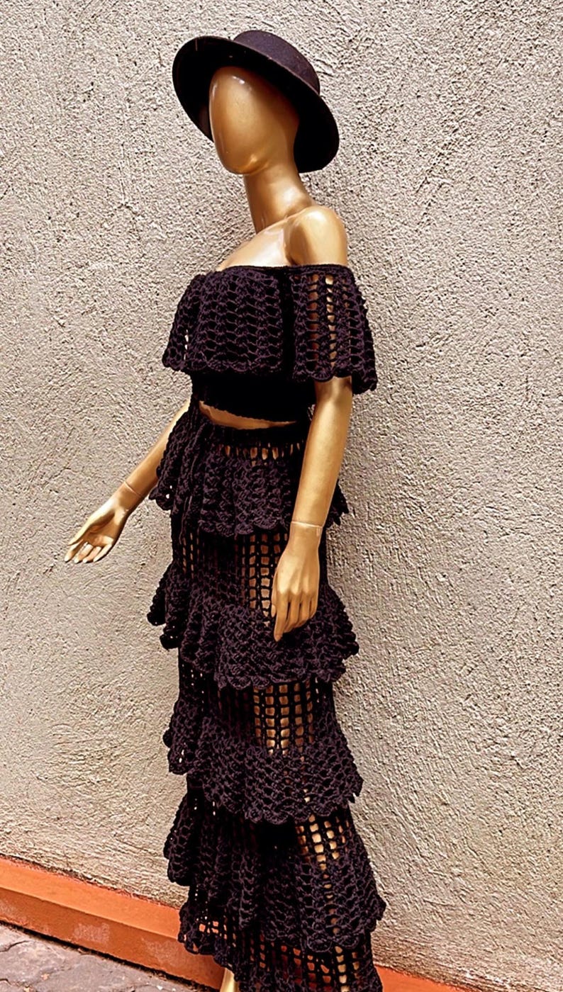 Peut inclure: Un ensemble deux pi&egrave;ces en crochet noir compos&eacute; d'un haut court &agrave; &eacute;paules d&eacute;nud&eacute;es avec des manches &agrave; volants et d'une jupe longue assortie &agrave; &eacute;tages. Le v&ecirc;tement pr&eacute;sente un motif en maille ouverte cr&eacute;ant un aspect textur&eacute; et superpos&eacute;.