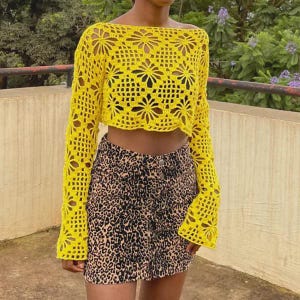 May include: A vibrant yellow crochet top with a floral design and long sleeves. The top is cropped and paired with a leopard print mini skirt. The outfit is set against a natural outdoor backdrop. The skirt has a button-up front.