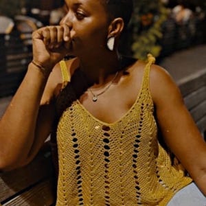 May include: Mustard yellow crochet tank top with a V-neck design. The top features a delicate open-weave pattern and small button details. The straps are a thicker crochet style. A gold necklace with a small pendant is visible.