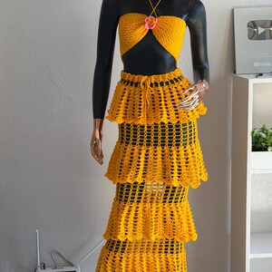 Peut inclure: Un ensemble deux pi&egrave;ces en crochet jaune vif compos&eacute; d'un haut court style dos nu avec une petite fleur rose au centre et une jupe longue &agrave; &eacute;tages assortie avec une taille &agrave; cordon, accompagn&eacute; d'un chapeau de paille &agrave; larges bords.
