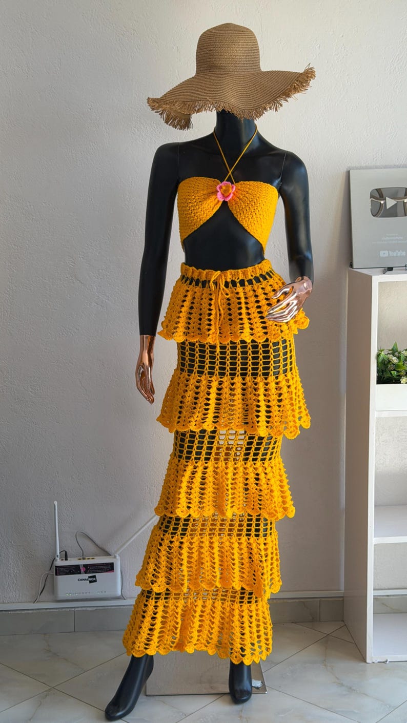 Peut inclure: Un ensemble deux pi&egrave;ces en crochet jaune vif compos&eacute; d'un haut court style dos nu avec une petite fleur rose au centre et une jupe longue &agrave; &eacute;tages assortie avec une taille &agrave; cordon, accompagn&eacute; d'un chapeau de paille &agrave; larges bords.