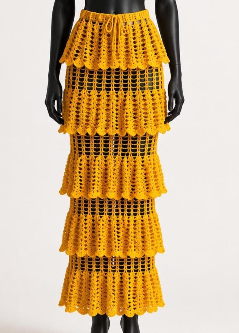 Peut inclure: Une jupe longue &agrave; &eacute;tages jaune vif pr&eacute;sentant un motif complexe en crochet ajour&eacute;. Le v&ecirc;tement poss&egrave;de une taille haute avec un cordon de serrage &agrave; la taille et quatre couches horizontales &agrave; volants distinctes descendant jusqu'au sol.