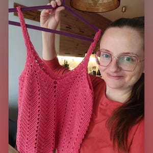 May include: A handmade, coral-colored crochet top with a V-neck and delicate stitch detailing. The top features thin straps and a fitted silhouette. The crochet pattern creates a textured, open design. The top is held up on a purple hanger.