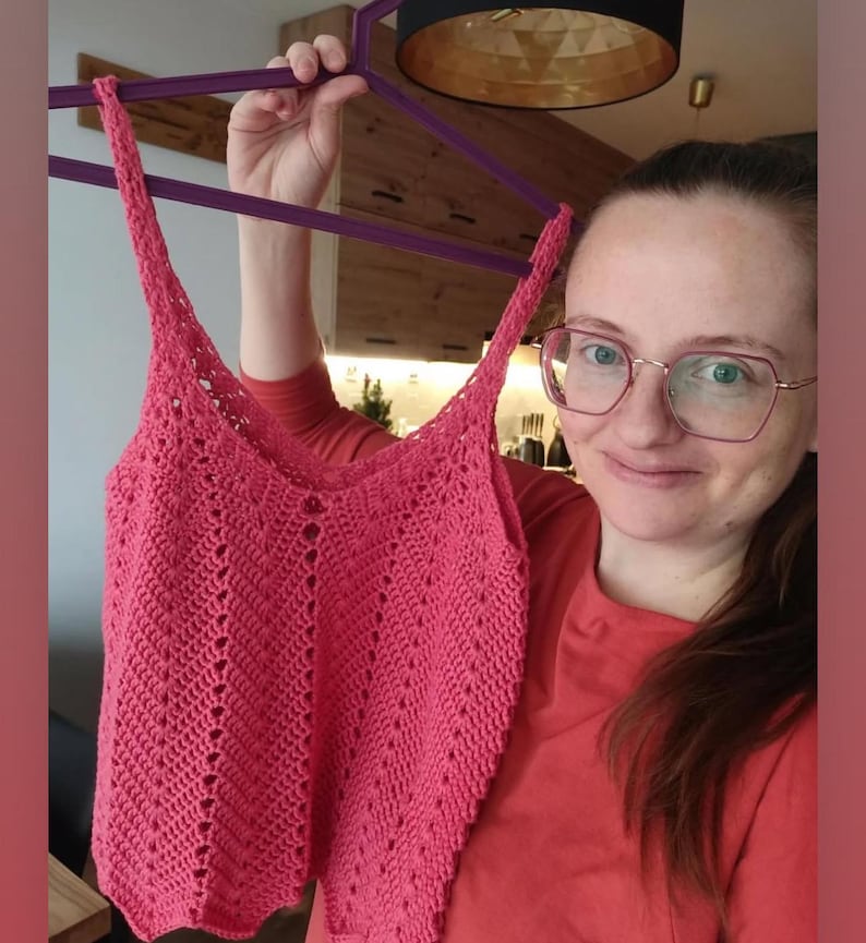 May include: A handmade, coral-colored crochet top with a V-neck and delicate stitch detailing. The top features thin straps and a fitted silhouette. The crochet pattern creates a textured, open design. The top is held up on a purple hanger.
