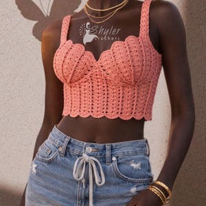 Op de afbeelding: Een koraalkleurige, gehaakte crop top met een geschulpte halslijn en bandjes. De top wordt gecombineerd met een spijkerbroekshort. De afbeelding toont ook gouden sieraden en een vlinderschaduw op de muur. De top is van Shyler Crochets.