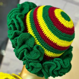 May include: A colourful crochet hat with a distinctive design. The hat's crown is striped with bands of red, yellow, and green. A ruffled green brim surrounds the crown, adding a decorative touch. The hat is likely intended for fashion or costume use.