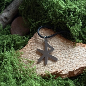 Könnte beinhalten: Ein silberfarbener Anhänger mit einem stilisierten Runen-Design hängt an einer schwarzen Kordel. Der Anhänger liegt auf einem Stück hellfarbenem Holz, umgeben von leuchtend grünem Moos. Der Hintergrund zeigt mehr grünes Moos und ein kleines Stück Holz.