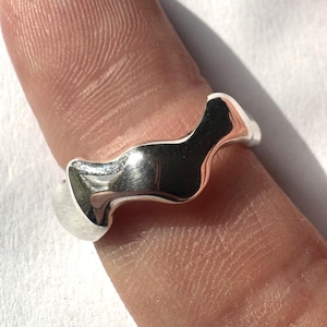 Könnte beinhalten: Ein polierter Silberring mit einem einzigartigen, fließenden Design. Der Ring hat eine glatte, reflektierende Oberfläche und eine wellenförmige, organische Form. Der Ring wird auf einem Finger gezeigt, um seine Form und Textur hervorzuheben.