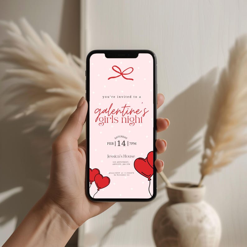 Op de afbeelding: Een smartphone toont een roze uitnodiging voor een "Galentine's Girls Night". De uitnodiging bevat rode hartballonnen, een strik en de datum "14 FEB | 19:00". De tekst luidt "Jessica's House" en andere evenementdetails.