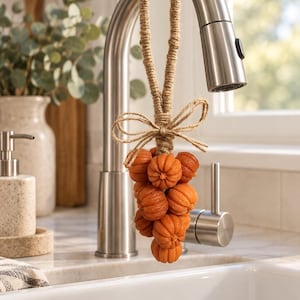 May include: A cluster of small, orange, pumpkin-shaped items suspended from a kitchen faucet by a thick, natural-colored jute rope. The rope is tied into a decorative bow at the top of the pumpkin cluster, which hangs vertically from the faucet spout.