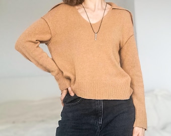 May include: A tan-colored sweater with a V-neck collar and long sleeves. The sweater is paired with black jeans. A silver pendant hangs from a black necklace. The model is standing with one hand on their hip.