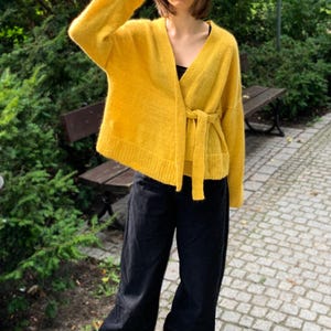 May include: A mustard yellow wrap cardigan with long sleeves and a tie belt. The cardigan is worn with black trousers. The image is set outdoors, with green bushes and a park bench in the background.