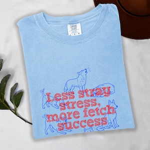 Peut inclure: T-shirt bleu clair à col rond. Le t-shirt présente un motif de chiens et le texte "Less stray, more fetch, success" en rouge. Le t-shirt est fait d'une matière douce.