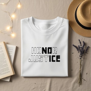 May include: A white t-shirt with the words "HONOR JUSTICE" printed in black. The shirt is folded neatly on a beige surface, next to a straw hat, a book, and a sprig of lavender.