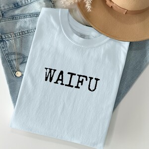 Könnte beinhalten: Hellblaues T-Shirt mit dem schwarzen Aufdruck "WAIFU". Das Shirt liegt gefaltet auf einer hellblauen Jeans. Ein beigefarbener Hut und eine goldene Halskette sind ebenfalls zu sehen.