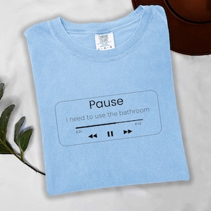 May include: Pale blue t-shirt with the text "Pause i need to use the bathroom" in a black rectangle, resembling a music player interface. The shirt also features a play/pause button and time indicators.
