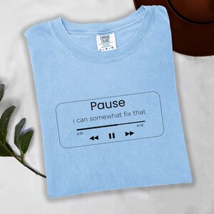 May include: Pale blue t-shirt with the words "Pause" and "i can somewhat fix that" in a black rectangle. The shirt also features a black music player graphic with play and pause buttons. The shirt is made of a soft material.