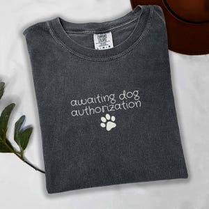 以下が含まれることがあります： 「awaiting dog authorization」の文字と白い足跡のグラフィックが入った、ダークグレーのComfort Colors Tシャツ。Tシャツは白い表面に折りたたまれています。背景には茶色の帽子が見えます。