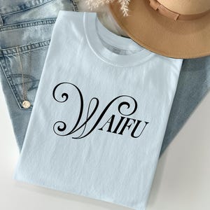 May include: A light blue t-shirt with the word "Waifu" in black script. The shirt is folded and placed on top of a pair of light blue jeans. A tan hat is visible in the background.