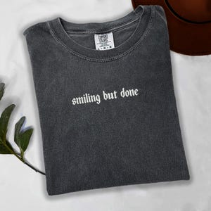 Puede incluir: Camiseta gris oscuro con la frase "smiling but done" en escritura blanca. La camiseta está doblada y tiene una etiqueta Comfort Colors. Un sombrero marrón es visible en el fondo.