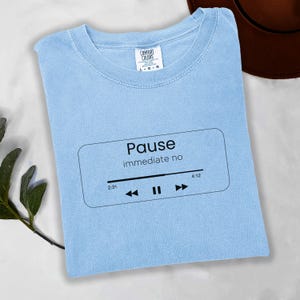May include: Pale blue t-shirt featuring the word "Pause" and "immediate no" in black, inside a rectangle. A black music player graphic is below the text. The shirt is a Comfort Colors brand.