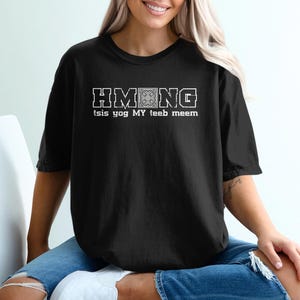 Puede incluir: Camiseta negra con texto blanco "HMONG" sobre las palabras "tsis yog MY teeb meem". La camiseta es de manga corta y cuello redondo.