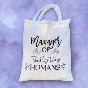 May include: A cream-coloured tote bag with the words "Manager of Thirty Tiny HUMANS" printed in black. The design includes illustrations of an apple, a mug, and hearts. The bag has two white handles.