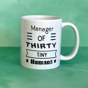 May include: White ceramic mug with a curved handle. The mug features black text that reads "Manager OF THIRTY TINY Humans" in a playful font. The mug is set against a green background.