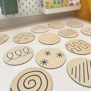 May include: A selection of wooden, oval discs featuring black line patterns. Designs include spirals, stars, dots, and geometric shapes. The discs are arranged on a white surface, possibly for a craft or educational purpose. Each disc is approximately 7 cm in diameter.