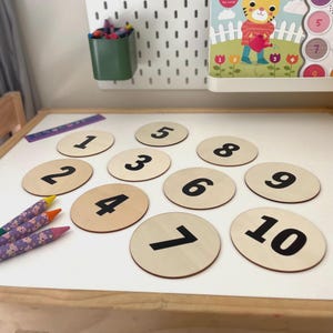 May include: Wooden number discs from 1 to 10 in black font on a light wood grain background. The discs are arranged on a white surface with crayons and a ruler nearby. A green container with crayons and a children's book are in the background.