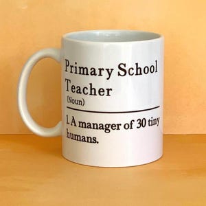 May include: White ceramic mug with a curved handle. The mug features black text that reads "Primary School Teacher (Noun) 1. A manager of 30 tiny humans."