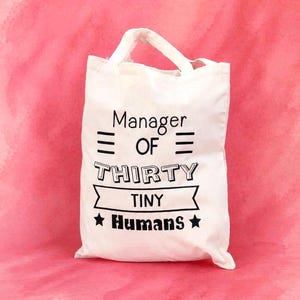 May include: Cream-coloured canvas tote bag featuring the text "Manager OF THIRTY TINY HUMANS" in black. The bag has two handles and is set against a pink background. The text is in a bold, playful font.