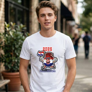 May include: White t-shirt with a cartoon soccer ball character wearing a hat and holding a Croatian flag. The shirt has the number "2026" above the character and the text "ŽIVJELA HRVATSKA" below.