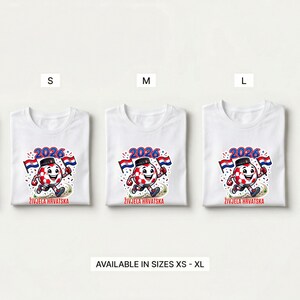 Pode incluir: T-shirts brancas com um design de personagem de bola de futebol. O design inclui o ano "2026", bandeiras croatas e o texto "ŽIVJELA HRVATSKA". Dispon&iacute;vel nos tamanhos XS a XL.